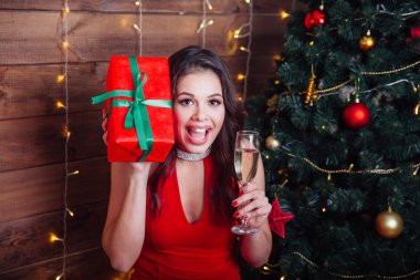 Kırmızı elbiseli zarif bir kadın elinde şampanya bardağı ve Noel ağacının yanında hediye kutusu tutuyor.