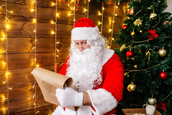 Santa Claus sitting at his room at home near Christmas tree and big sack and reading Christmas letter or wish list — 스톡 사진