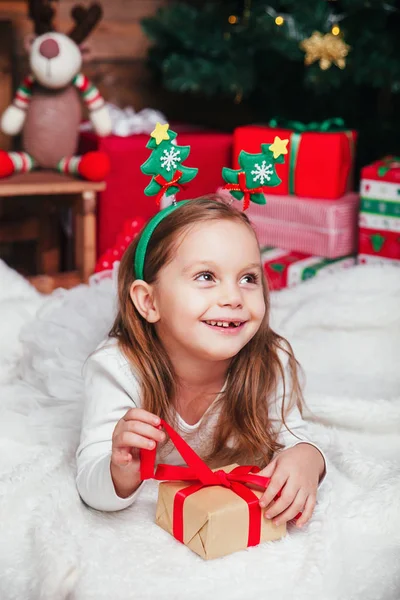 Счастливая маленькая улыбающаяся девочка с рождественской подарочной коробкой, лежащей рядом с елкой — стоковое фото
