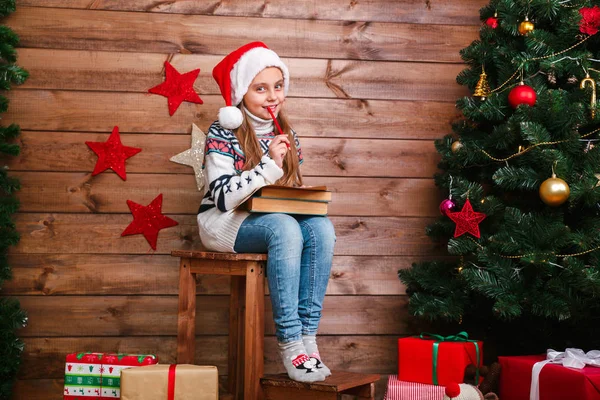 Muchacha divertida en Santa sombrero escribe carta a Santa cerca del árbol de Navidad — Foto de Stock