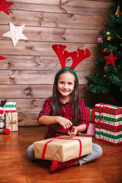 Feliz niña divertida en cuernos de ciervo paquete de regalos cerca del árbol de Navidad —  Fotos de Stock