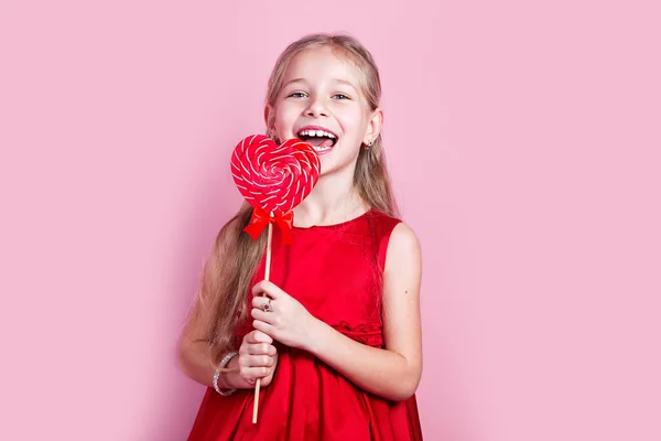 Μικρό χαρούμενο κορίτσι με κόκκινο φόρεμα με ένα γλειφιτζούρι σε σχήμα καρδιάς — Φωτογραφία Αρχείου