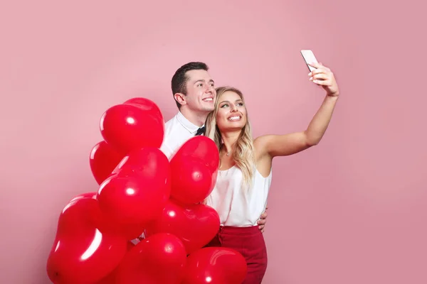 Schönes junges Paar macht Selfie auf Handy und steht mit roten Luftballons auf rosa Hintergrund — Stockfoto