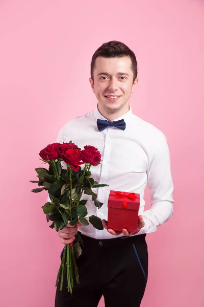 Bello uomo spillante che tiene fiori e scatola regalo su sfondo rosa . — Foto Stock