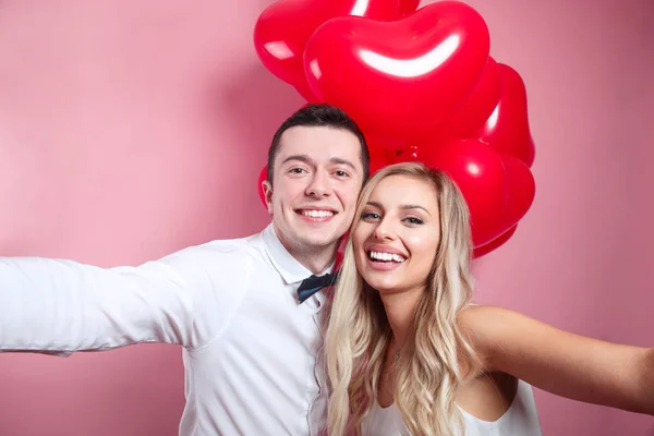 Beau jeune couple prenant selfie sur téléphone portable et debout avec des ballons rouges sur fond rose — Photo