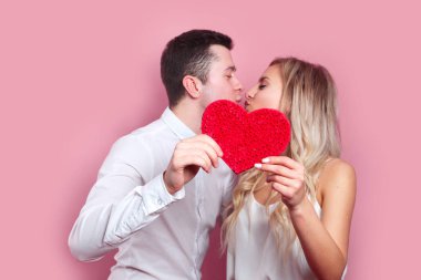 Genç çift pembe arka planda kağıt kalbin arkasında öpüşüyor.