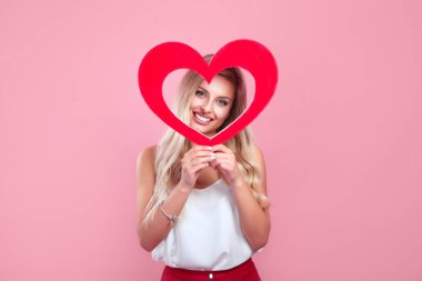 Güzel, gülümseyen bir kadın pembe arka planda kırmızı bir kalp tutuyor..
