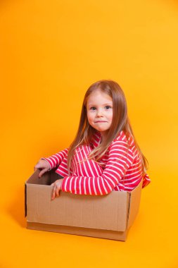 Mutlu küçük kız sarı arka planda karton bir kutuda oturuyor.