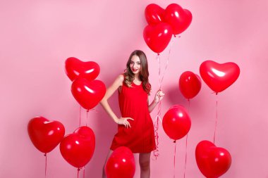 Pembe arka planda kalp şeklinde kırmızı balonları olan kırmızı elbiseli güzel, zarif genç bir kadın..