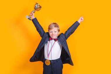 Sarı arka planda altın bir kupa ve altın madalya taşıyan neşeli çocuk.