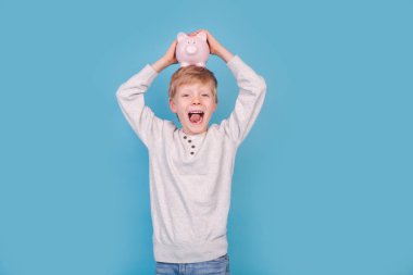 Mavi arka planda kumbarası olan heyecanlı küçük çocuk.