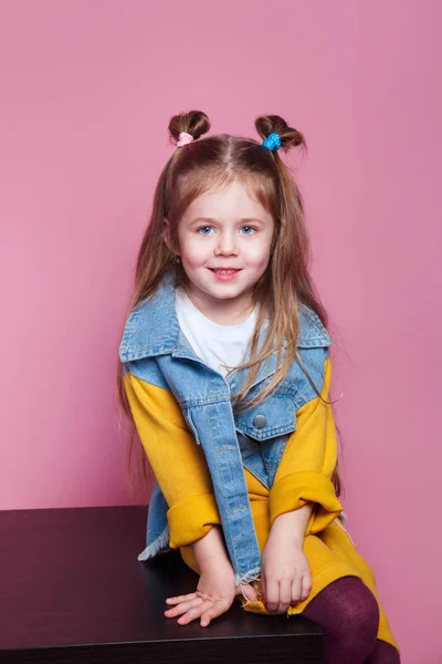 Beautiful stylish little girl in jeans clothes sitting and smiling on pink background — Φωτογραφία Αρχείου