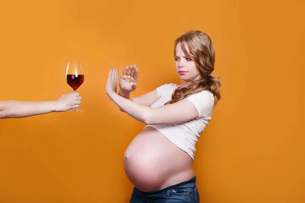 Pregnant woman refuses to drink wine, making stop gesture to glass on yellow background — Zdjęcie stockowe