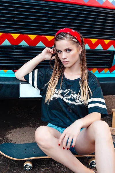 Beautiful smiling girl, with many pigtails wearing in shirt and shorts sitting on skateboard on the street — Stock Photo, Image