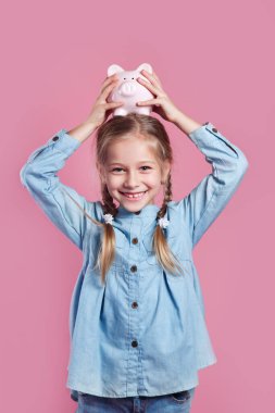 Mavi gömlekli şirin küçük kız pembe arka planda domuzcuk kutusunu sallıyor.