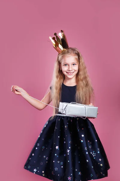 Χαριτωμένο χαμογελαστό κοριτσάκι με φόρεμα πριγκίπισσα και το στέμμα κρατώντας το παρόν κουτί σε ροζ φόντο — Φωτογραφία Αρχείου