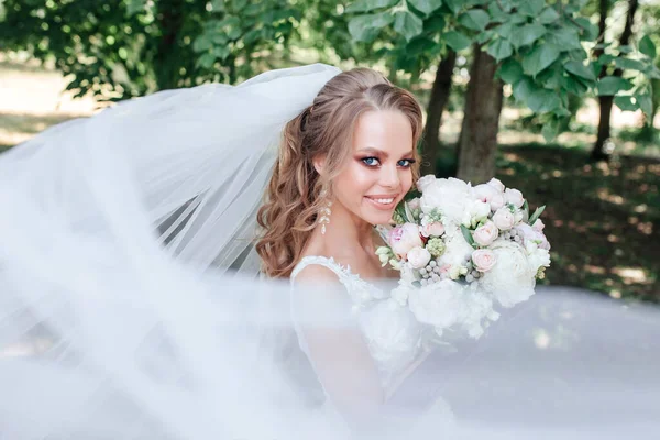 Hermosa novia con ramo de flores de boda, mujer atractiva en vestido de novia con velo volador en el parque — Foto de Stock