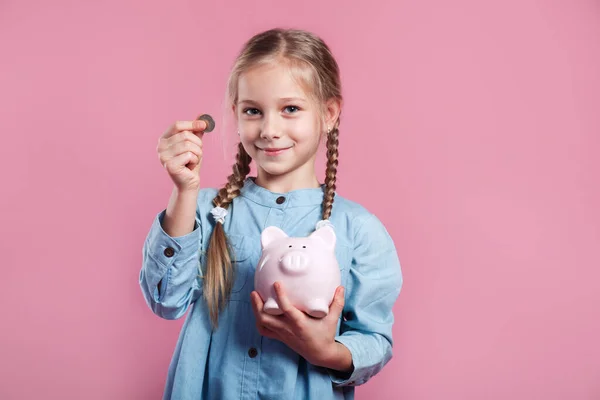 Маленькая девочка копит деньги в копилке на розовом фоне — стоковое фото