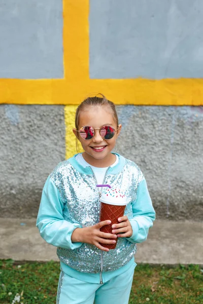 Маленька стильна дівчинка в сонцезахисних окулярах п'є молочний коктейль на міському фоні — стокове фото