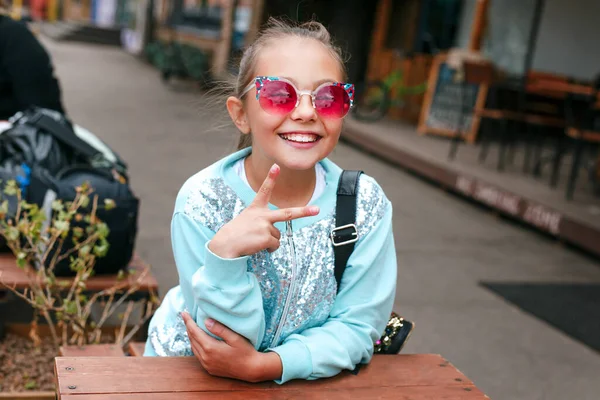 Küçük kız güneş gözlüğü ve sırt çantası takıyor şehrin arka planında oturuyor. — Stok fotoğraf