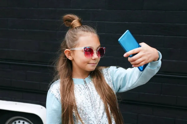 Güneş gözlüklü küçük güzel bir kız akıllı bir telefonla selfie çekiyor ve dışarıda gülümsüyor. — Stok fotoğraf