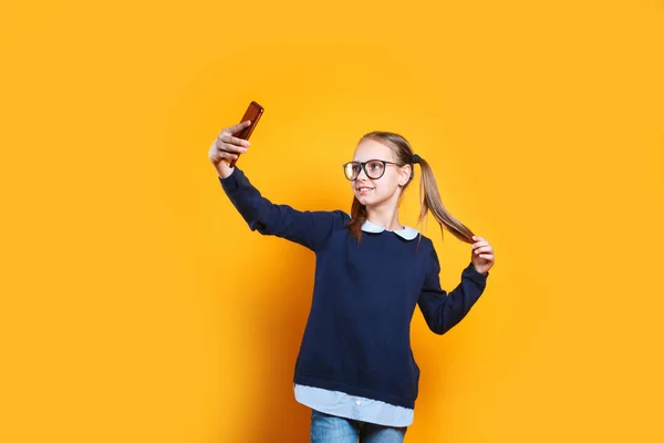 안경을 쓴 쾌활 한 어린 소녀가 노란 배경에서 셀카를 찍는 휴대 전화를 사용하는 모습 — 스톡 사진