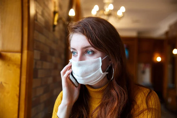 Mujer europea con el pelo rojo en máscara médica protectora hablando por teléfono en la cafetería — Foto de Stock