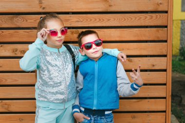 Şehirdeki moda çocukları ahşap bir duvarda duruyorlar. Güneş gözlüklü kız ve oğlan sokakta dikiliyorlar.