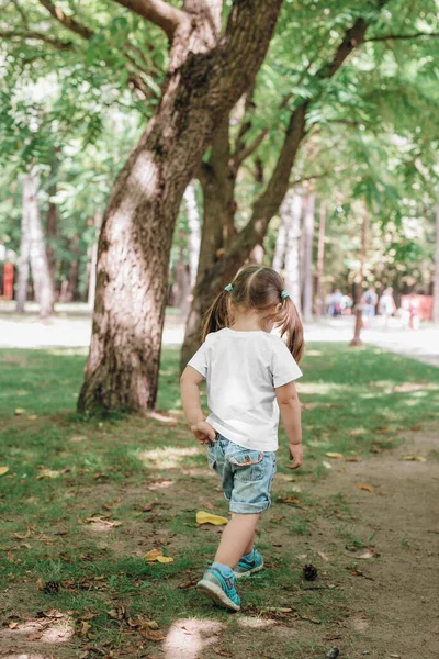 Tillbaka utsikt över liten flicka i blank t-shirt promenader i parken. Mockup för design — Stockfoto