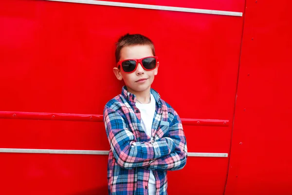 Portré kisfiú kockás inget és napszemüveget visel a városban a vörös háttér felett a városban — Stock Fotó
