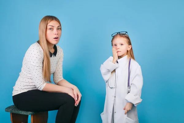 Cute little girl as doctor playing with happy mom on blue studio background