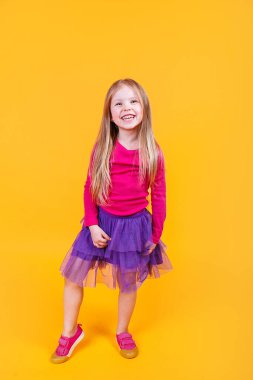 Sarı arka planda duran pembe tişörtlü ve mor etekli mutlu ve sevimli, komik, modaya uygun küçük kız.