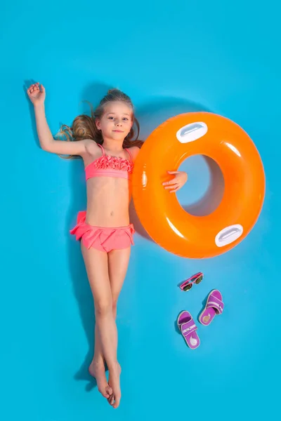 Mavi arka planda şişme daire ve terliklerle yerde yatan küçük bir kızın en üst görüntüsü.