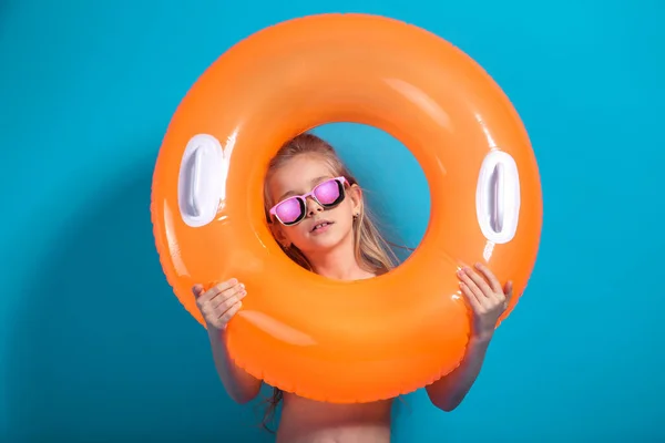 Happy baby girl w stroju kąpielowym i okulary przeciwsłoneczne z pomarańczowym pierścieniem pływackim na kolorowe niebieskie tło — Zdjęcie stockowe