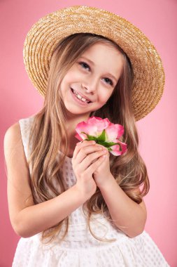 Yaz kıyafetleri ve hasır şapkalı çekici kız pembe stüdyo arka planında elinde bir çiçek tutuyor..