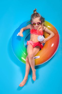 Mayo giymiş küçük bir kız, sarı stüdyo arka planında dondurma olan şişirilebilir renkli bir yüzüğün üzerinde yatıyor..