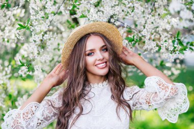 Hasır şapkalı, kıvırcık uzun saçlı, genç ve çekici bir kadın bahar günü çiçek açan bahçenin tadını çıkarıyor..