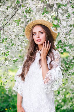 Uzun kıvırcık saçlı, şık hasır şapkalı yeşil çiçekli bahçede yürüyen genç bir kadın..