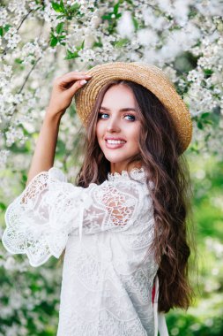 Kıvırcık uzun saçlı, saman şapkalı, güneşli bahar gününde çiçek açan bahçede poz veren genç ve çekici bir kadın..