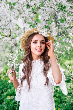 Kıvırcık uzun saçlı, saman şapkalı, güneşli bahar gününde çiçek açan bahçede poz veren genç ve çekici bir kadın..