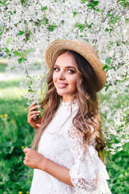 Hasır şapkalı, kıvırcık uzun saçlı, genç ve çekici bir kadın güneşli bahar gününde çiçek açan bahçenin tadını çıkarıyor..