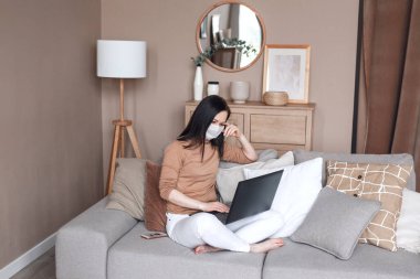 Koronavirüs yüzünden karantinada olan bir kadın koruyucu maske takıyor ve merkez bürodaki kanepede çalışıyor.