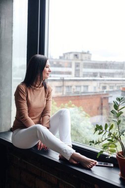 Camdan bakan, şehir manzaralı, pencere kenarında oturan, rahatlatıcı genç bir kadın.
