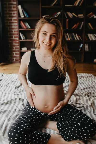 Atractiva mujer embarazada está sentada en la cama y sosteniendo su campana —  Fotos de Stock
