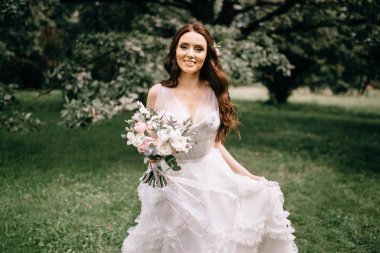 Çiçekli güzel gelin orman arka planında duruyor. Dışarıda zarif elbiseler içinde güzel bir gelin. Güneşli, ağaçlık alanda parkta genç bir gelinin portresi..