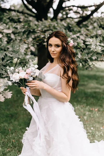 Schöne Braut mit Blumenstrauß steht auf Waldhintergrund. schöne Braut in zartem Kleid im Freien. Nahaufnahme Porträt der jungen Braut im Park bei sonnigem Wetter Waldgebiet. — Stockfoto