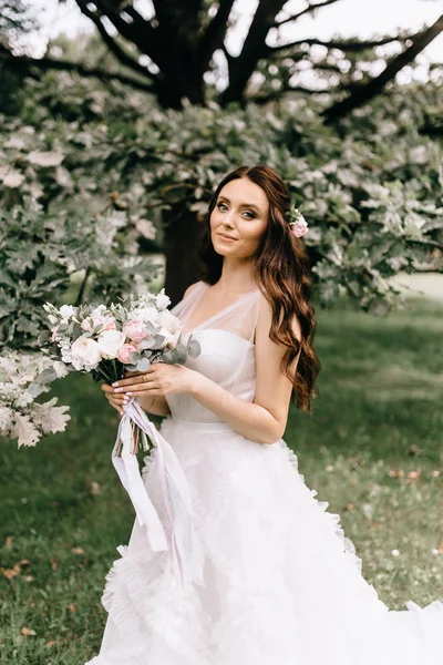 Όμορφη νύφη με λουλούδια μπουκέτο στέκεται στο δάσος φόντο. όμορφη νύφη σε λεπτή φόρεμα σε εξωτερικούς χώρους. Κοντινό πλάνο πορτρέτο της νεαρής νύφης στο πάρκο στην ηλιόλουστη δασώδη περιοχή. — Φωτογραφία Αρχείου