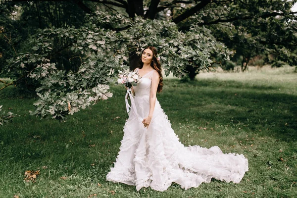 Όμορφη νύφη με λουλούδια μπουκέτο στέκεται στο δάσος φόντο. όμορφη νύφη σε λεπτή φόρεμα σε εξωτερικούς χώρους. Κοντινό πλάνο πορτρέτο της νεαρής νύφης στο πάρκο στην ηλιόλουστη δασώδη περιοχή. — Φωτογραφία Αρχείου