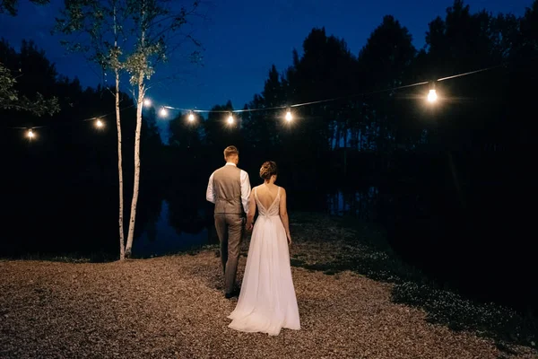 Fashionable newlyweds stand at night against the backdrop of chic decorations and flowers,electric bulbs and garlands,in the backlight. Night shooting of bride and groom.Floristics.
