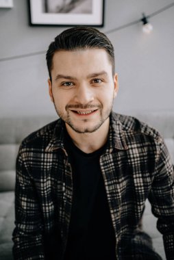 Çekici, gülümseyen, sakallı genç bir adamın portresi. Günlük giysiler giymiş. Oturma odasında bir kanepede oturuyor.